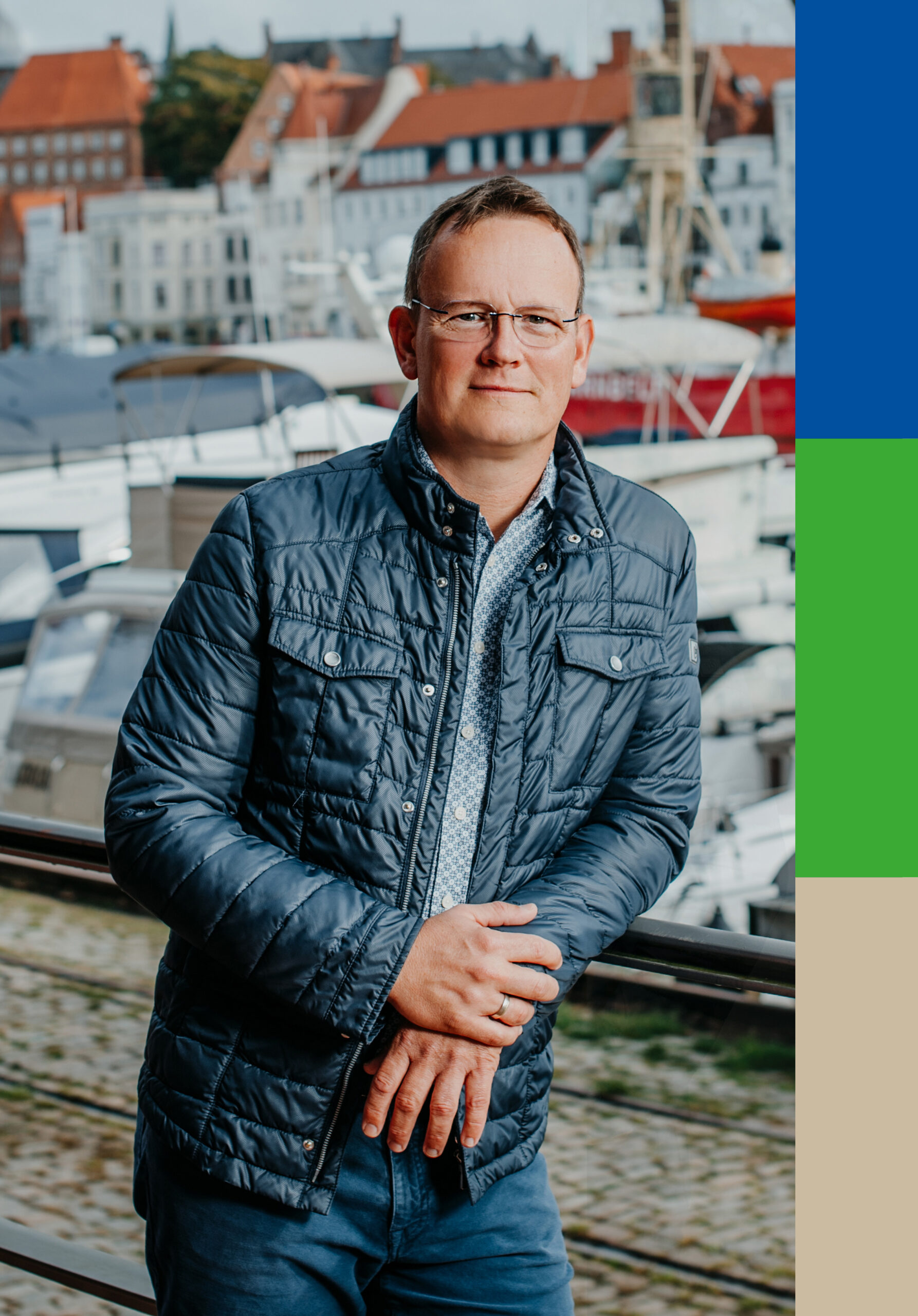 Porträt von Frank Belau, Geschäftsführer von belau-betreuung.de, mit Brille und dunkelblauer Jacke vor einer maritimen Altstadtkulisse.