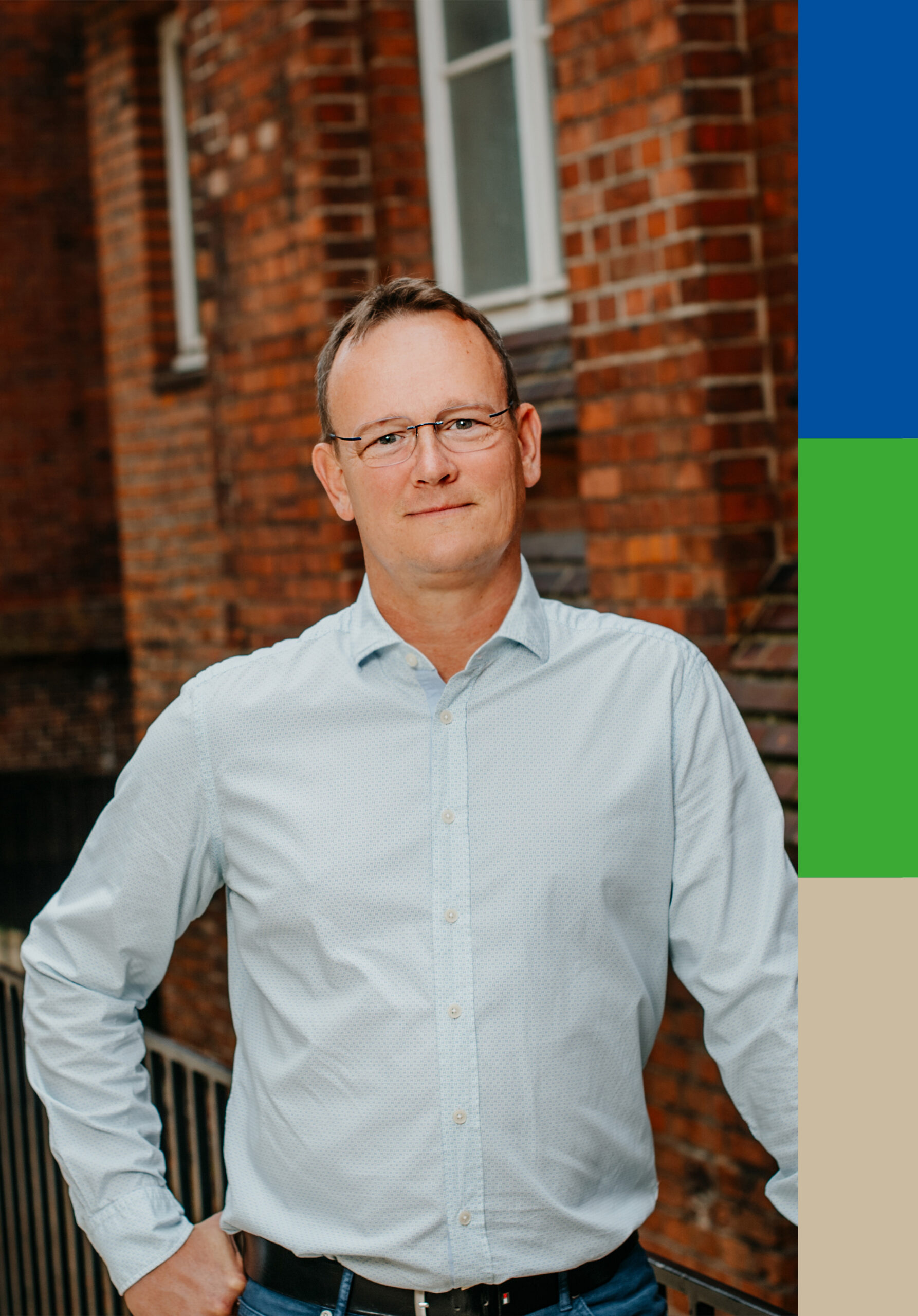 Porträt Frank Belau, Geschäftsführer, im hellblauen Hemd, lächelnd vor einer roten Backsteinwand.